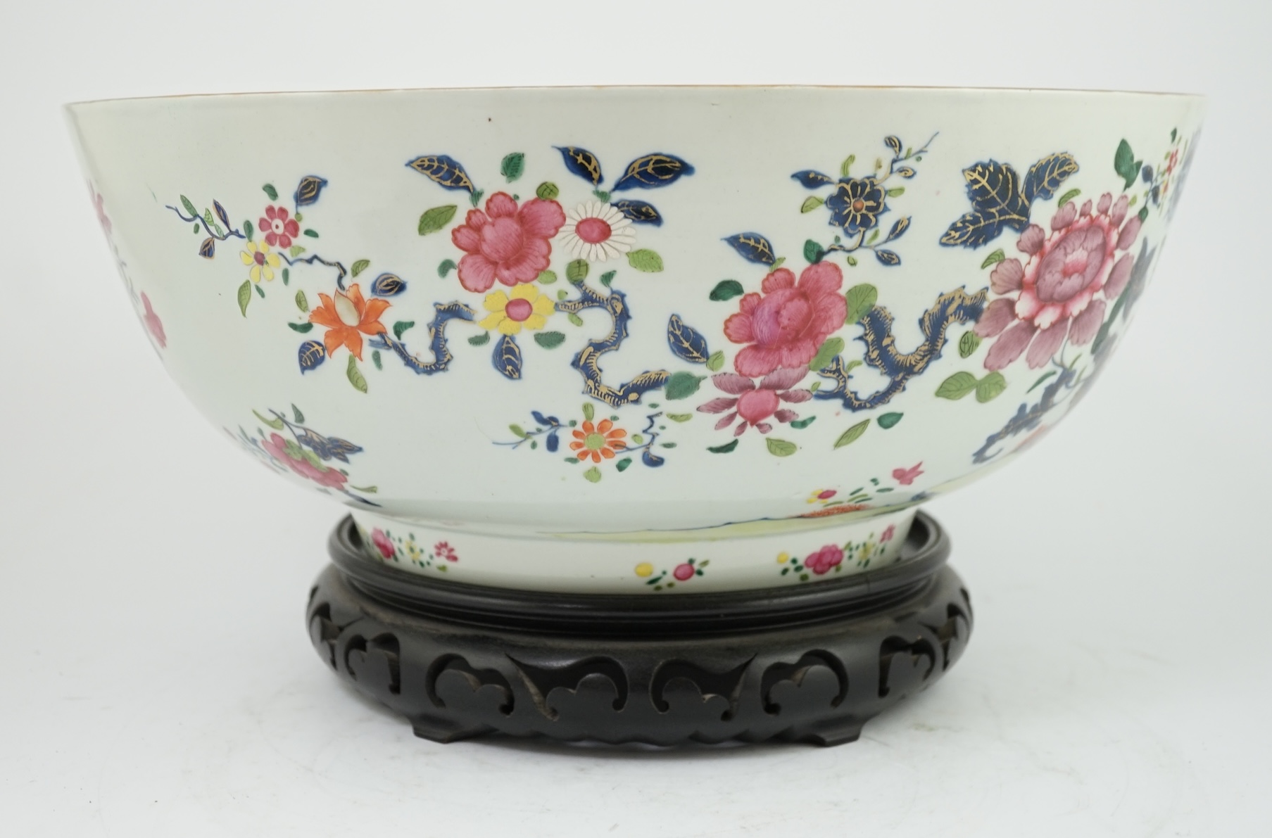 A large Chinese famille rose punch bowl, Qianlong period, the exterior painted with flowers in a fenced garden, the interior with a flower bouquet within a garland of flowers, 40.5cm diameter, wood stand. Condition - som